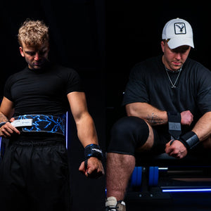 Two men standing side by side with lifting belts, knee sleeves, and wrist wraps in gym