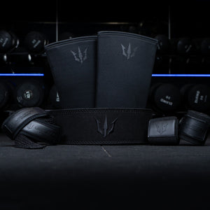 Black Unlisted Strength bundle consisting of lever belt, knee sleeves, wrist wraps, and lifting straps in front of dumbbells in a gym