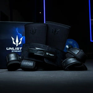 Black Unlisted Strength bundle consisting of lever belt, knee sleeves, wrist wraps, and lifting straps assembled in front of an Unlisted Strength box in a gym