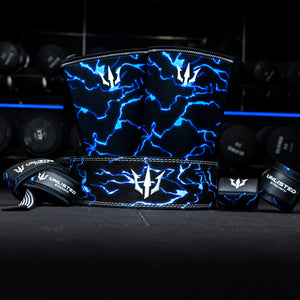 Blue Unlisted Strength bundle consisting of lever belt, knee sleeves, wrist wraps, and lifting straps in front of dumbbells in a gym